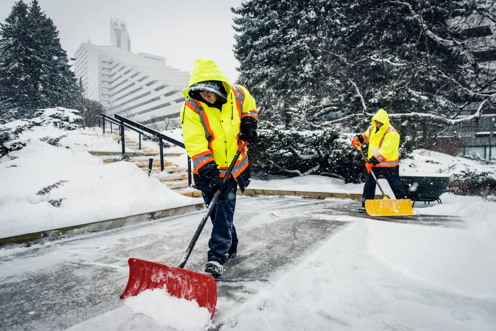 Deux employés en dossard haute visibilité déneigent à la pelle pendant une tempête
